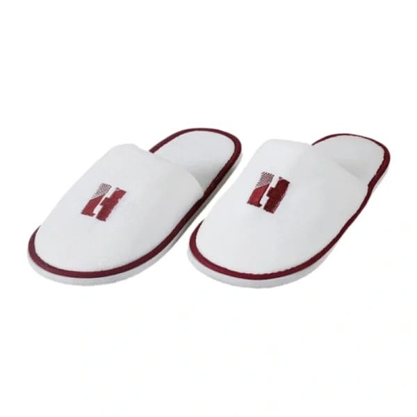 A white Hotel Velour Slippers neatly placed on a hotel room floor next to a bed, symbolizing a welcoming guest amenity.