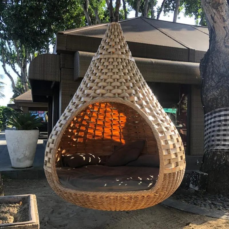 A couple relaxing together in a large, round Hanging swing bed rattan bird nest bed on a private hotel villa terrace, overlooking a tropical sunset.