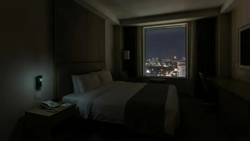 A Hotel Emergency torch charging on a wall mount next to a bed in a dark hotel guest room, providing a reassuring green glow during a nighttime power outage.