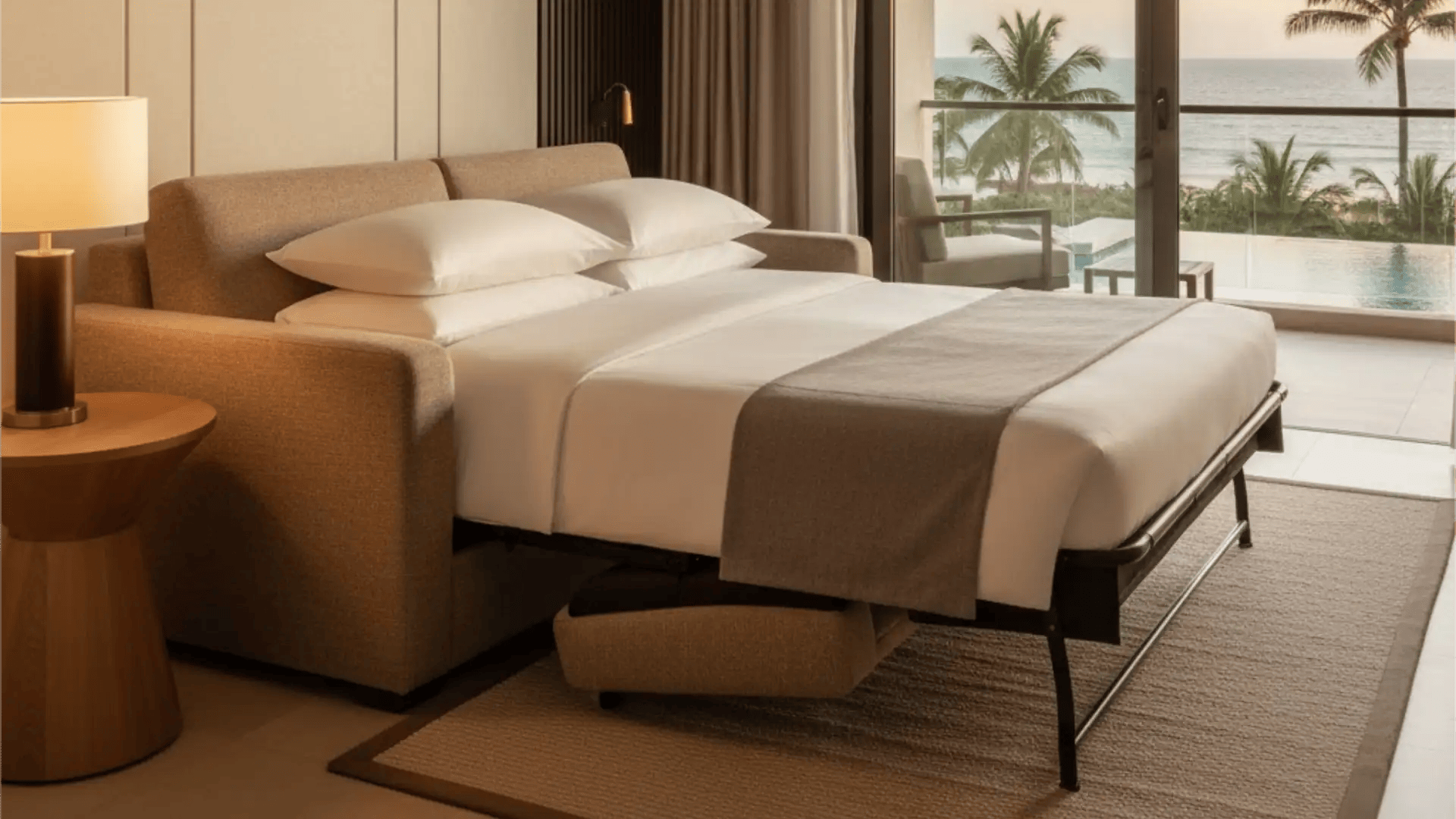 A modern beige commercial grade hotel sofa bed fully unfolded into a comfortable queen size bed in a luxury ocean view resort suite.