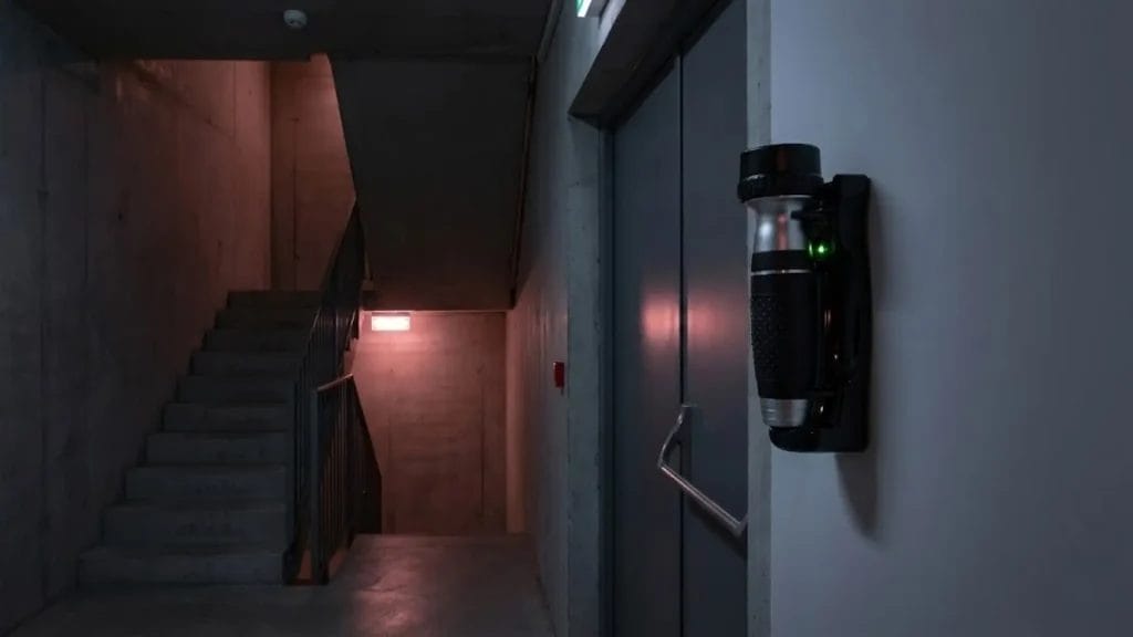 A wall-mounted Hotel Emergency torch with a charging base, featuring a green ready light, installed in a dimly lit hotel emergency stairwell next to a fire door.