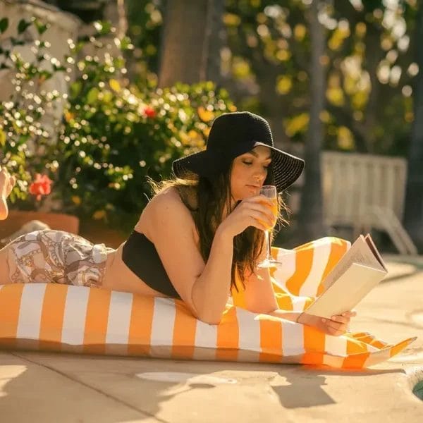 A woman wearing a sun hat and sipping a drink while lying on an orange and white striped bean bag lounger by the pool.